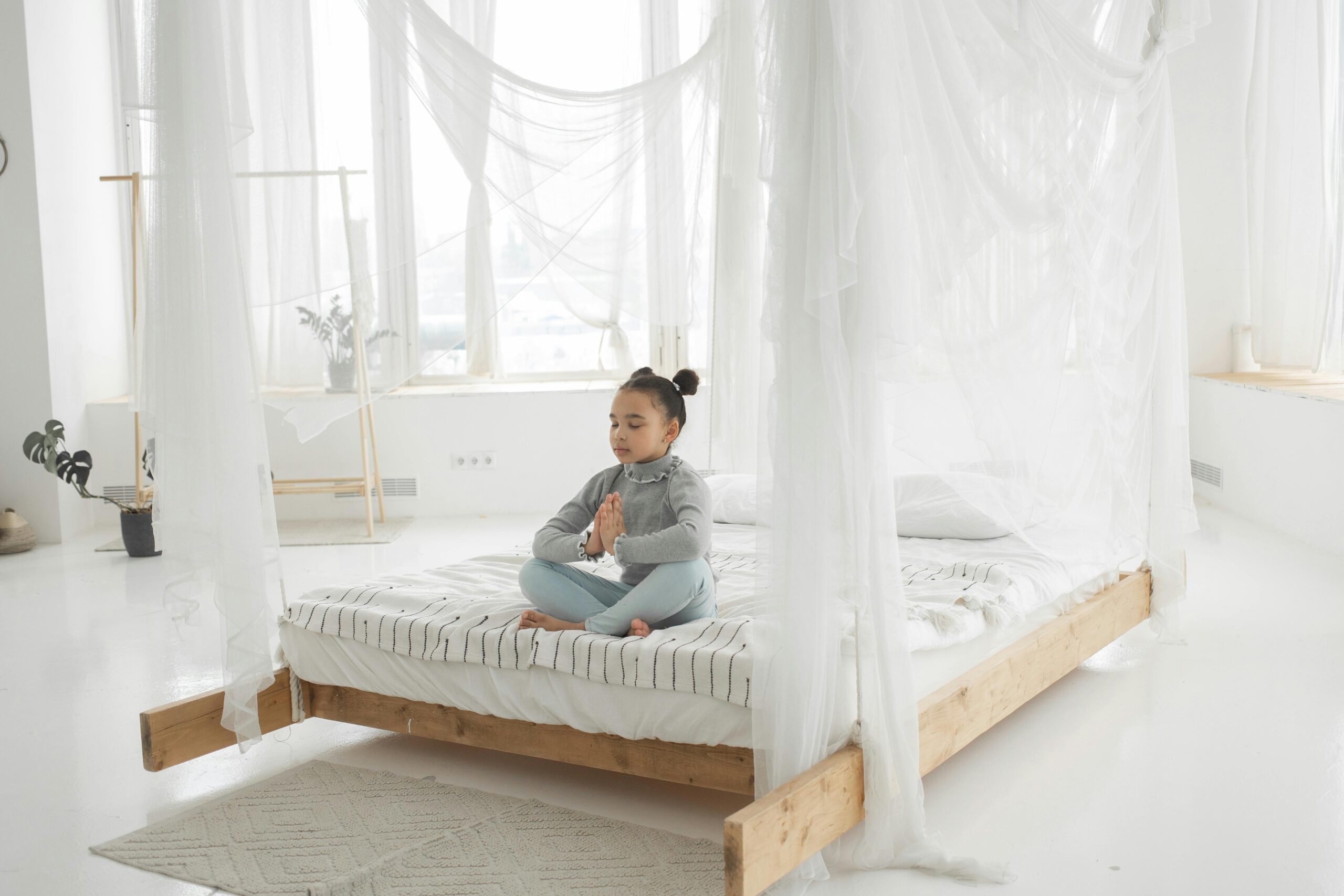 Full length of peaceful barefooted little ethnic kid in casual clothes meditating with closed eyes sitting on comfortable bed in Lotus pose in daylight